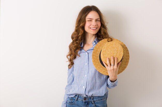 Retrato de joven mujer bonita con sombrero de paja jeans camisa de algodón azul posando en la pared blanca