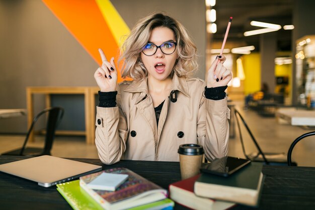 Retrato de joven mujer bonita teniendo una idea, sentado a la mesa en gabardina trabajando en la computadora portátil en la oficina de trabajo compartido, con gafas, ocupado, pensando, problema