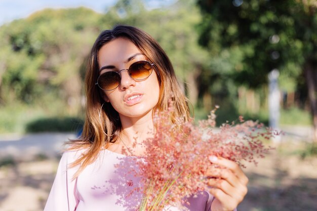 Retrato de joven mujer bronceada caucásica en romántico vestido rosa aretes redondos pulsera de plata y gafas de sol con flores silvestres