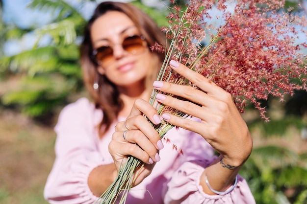 Foto gratuita retrato de joven mujer bronceada caucásica en romántico vestido rosa aretes redondos pulsera de plata y gafas de sol con flores silvestres