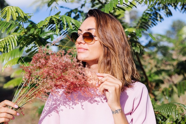 Retrato de joven mujer bronceada caucásica en romántico vestido rosa aretes redondos pulsera de plata y gafas de sol con flores silvestres