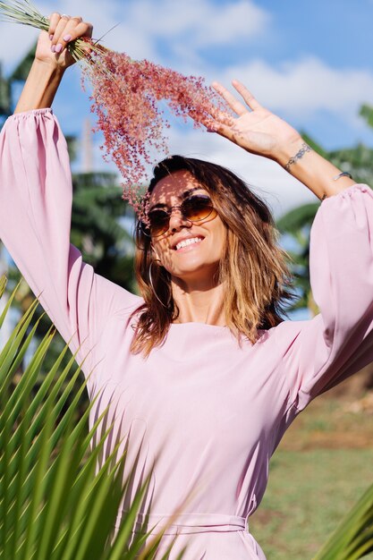 Retrato de joven mujer bronceada caucásica en romántico vestido rosa aretes redondos pulsera de plata y gafas de sol con flores silvestres