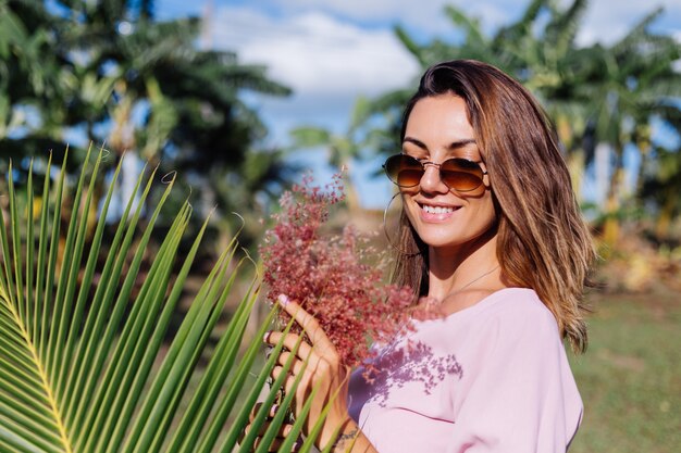 Retrato de joven mujer bronceada caucásica en romántico vestido rosa aretes redondos pulsera de plata y gafas de sol con flores silvestres