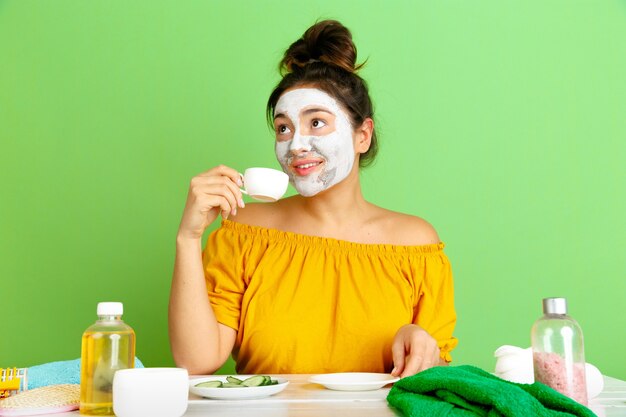 Retrato de joven mujer caucásica en el día de la belleza, rutina de cuidado de la piel y el cabello. Modelo femenino tomando café, té mientras se aplica la mascarilla facial. Cuidado corporal y facial, belleza natural y concepto de cosmética.