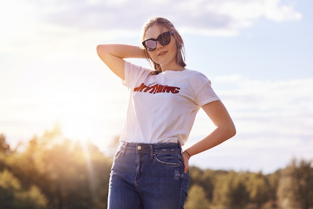 Retrato de joven mujer elegante con ropa casual y tonos, disfruta del horario de verano, posa en un cielo despejado, tiene un peinado moderno. Adolescente admira la hermosa naturaleza. Gente, estilo, amistad