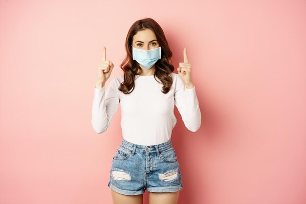 Retrato de una joven mujer elegante con ropa informal de verano que usa una máscara médica en la cara señalando con el dedo...
