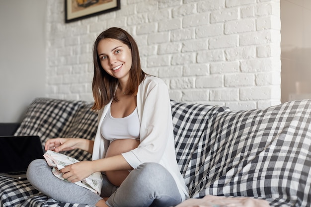 Retrato de joven mujer embarazada preparando ropa para su bebé recién nacido sonriendo sentado en un acogedor sofá en casa. Concepto de embarazo y expectativas.