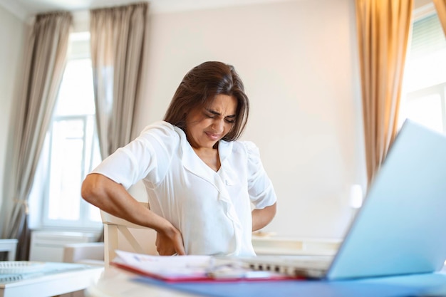 Retrato de una joven mujer estresada sentada en el escritorio de la oficina en casa frente a una laptop tocando la espalda dolorida con expresión de dolor que sufre de dolor de espalda después de trabajar en una laptop