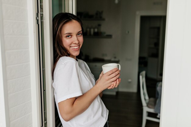 Retrato de joven mujer europea con cabello oscuro y piel sana sonriendo con café de la mañana en la moderna cocina ligera.