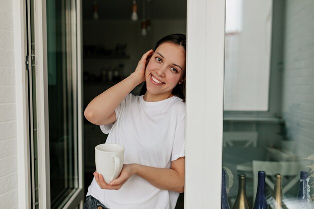 Retrato de joven mujer europea con cabello oscuro y piel sana sonriendo con café de la mañana en la moderna cocina ligera.