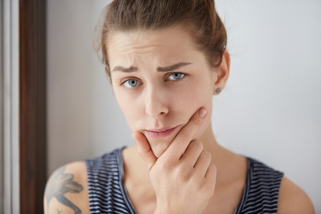 Retrato de joven mujer europea tatuada mostrando sospecha con cejas oscuras fruncidas. Hermosa chica morena en top pelado sosteniendo su barbilla con el pulgar y el dedo índice atascado en duda.