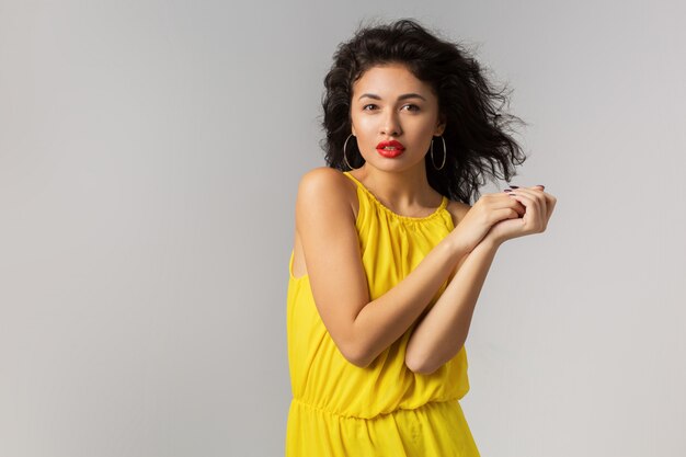 Retrato de joven mujer exótica atractiva en vestido amarillo elegante, labios rojos, aspecto natural, piel pura, sonriente, aislado, estilo de temporada de verano, tendencia de moda, viento que sopla en el pelo rizado