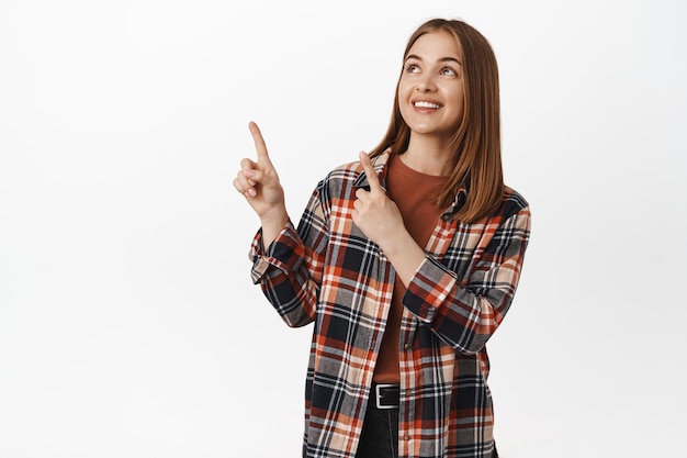 Retrato de una joven mujer feliz, una chica que apunta a la esquina superior izquierda, mirando la pancarta del logo, sonriendo alegre, de pie con un traje casual de gen-z, fondo blanco. copia espacio