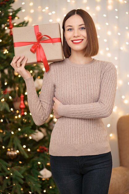 Retrato de joven mujer feliz con labios rojos mirando a la cámara sosteniendo una caja de regalo envuelta