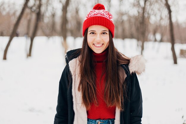 Retrato de joven mujer feliz muy sonriente en suéter rojo y gorro de punto con abrigo de invierno, caminando en el parque en la nieve, ropa de abrigo