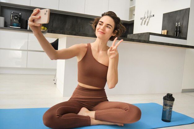 Retrato de una joven mujer fitness sentada en casa sobre una alfombra de goma graba un video en un teléfono inteligente haciendo trabajo