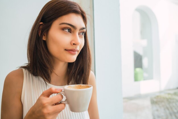 Retrato de joven mujer latina disfrutando y bebiendo una taza de café en la cafetería.