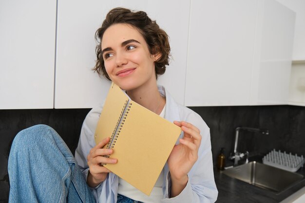 Retrato de una joven mujer moderna sentada en la cocina hojeando páginas leyendo un libro de diario sonriendo