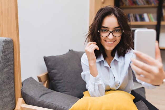 Retrato joven mujer morena bastante sonriente en gafas negras relajante en el sofá en el apartamento moderno. Usando el teléfono, haciendo selfie, sonriendo, estado de ánimo alegre.