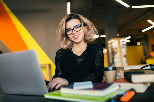 Retrato de joven mujer muy sonriente sentada a la mesa en camisa negra trabajando en la computadora portátil en la oficina de trabajo conjunto, con gafas