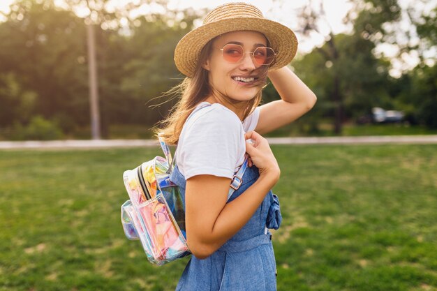 Retrato de joven mujer muy sonriente con sombrero de paja y gafas de sol rosas caminando en el parque, estilo de moda de verano, traje colorido hipster