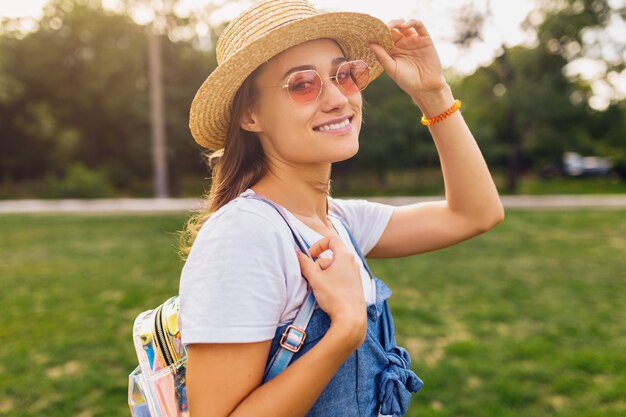 Retrato de joven mujer muy sonriente con sombrero de paja y gafas de sol rosas caminando en el parque, estilo de moda de verano, traje colorido hipster