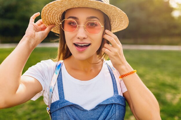 Retrato de joven mujer muy sonriente con sombrero de paja y gafas de sol rosas caminando en el parque, estilo de moda de verano, traje colorido hipster