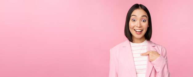 Retrato de una joven mujer de negocios asiática con una expresión sorprendida de la cara emocionada apuntándose con el dedo a sí misma de pie con traje sobre fondo rosa