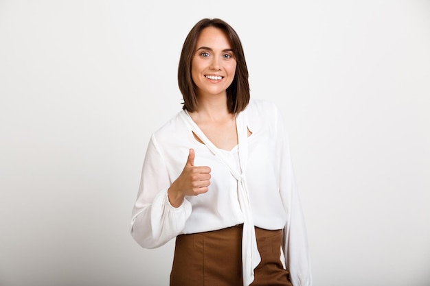 Foto gratuita retrato de joven mujer de negocios exitosa sonriendo, mostrando el pulgar hacia arriba, mirando al frente, sobre la pared blanca.