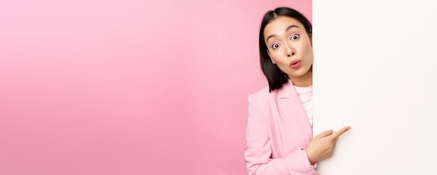 Retrato de una joven mujer de negocios japonesa dama corporativa en traje apuntando a la pared con un gráfico que muestra un diagrama o un anuncio en el espacio vacío de la copia fondo rosa