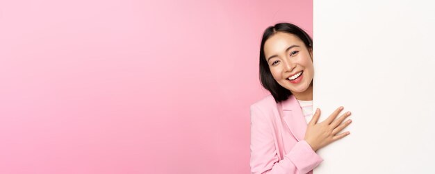 Retrato de una joven mujer de negocios japonesa dama corporativa en traje apuntando a la pared con un gráfico que muestra un diagrama o un anuncio en el espacio vacío de la copia fondo rosa