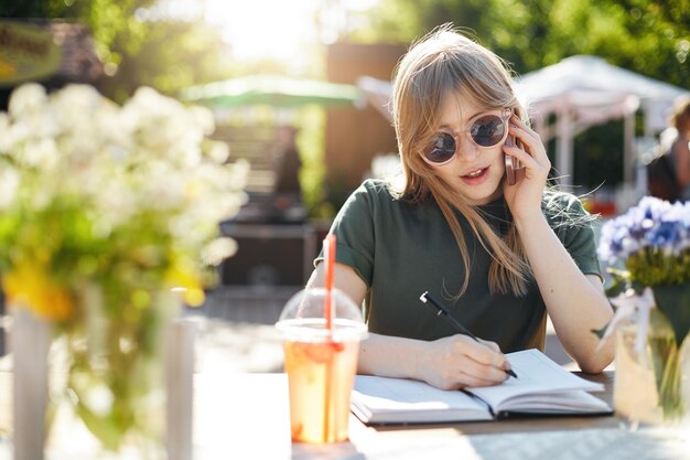 Retrato de una joven mujer de negocios o estudiante escribiendo sus planes en un bloc de notas hablando en un teléfono inteligente con gafas durante un almuerzo en el parque en un día soleado de verano Copiar espacio