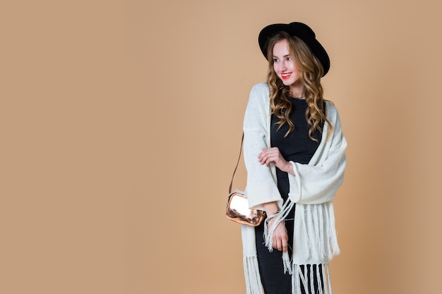 Retrato de joven mujer rubia elegante con sombrero de lana negro vistiendo poncho de flecos blancos de gran tamaño con vestido largo gris