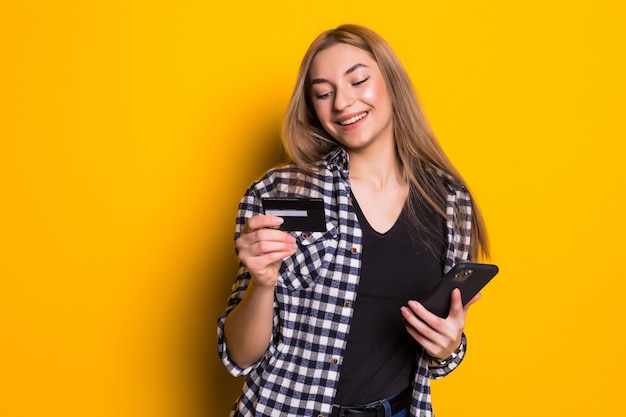 Retrato de una joven mujer rubia feliz mostrando una tarjeta de crédito de plástico mientras usa un teléfono móvil aislado sobre una pared amarilla