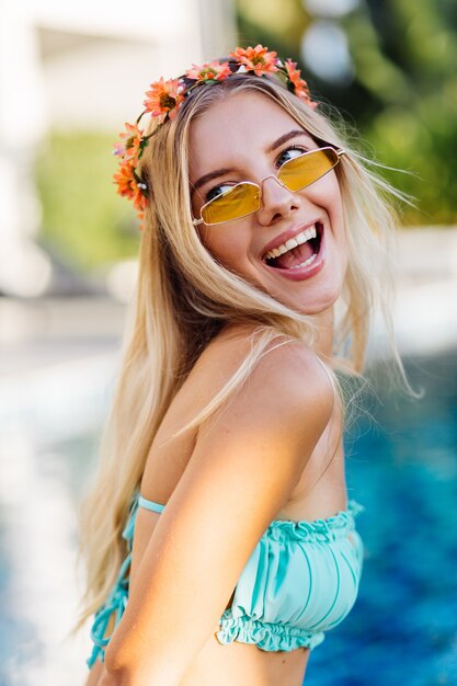 Retrato de joven mujer rubia feliz de pelo largo en bikini azul y corona de flores en la cabeza.