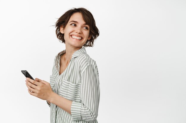 Retrato de joven mujer sonriente sosteniendo teléfono inteligente, gire la cabeza en el espacio de la copia, charlando celular, de pie en blanco. Copia espacio