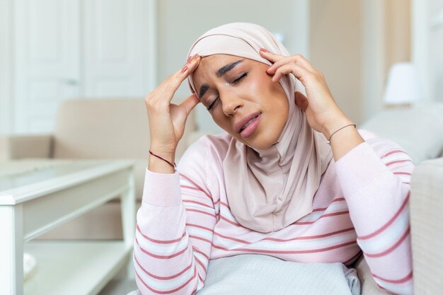 Retrato de una joven musulmana árabe sentada en el sofá de su casa con dolor de cabeza y dolor