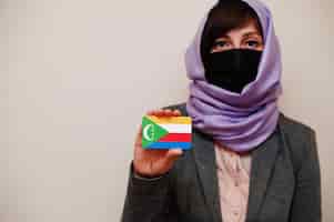 Foto gratuita retrato de una joven musulmana con ropa formal que protege la máscara facial y el pañuelo de la cabeza del hiyab sostiene la tarjeta de la bandera de las comoras contra un fondo aislado concepto de país del coronavirus