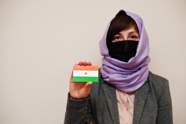 Retrato de una joven musulmana con ropa formal que protege la máscara facial y el pañuelo para la cabeza del hiyab sostiene la tarjeta de la bandera de Níger contra un fondo aislado Concepto de país del coronavirus