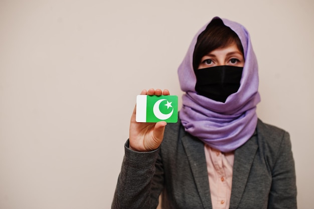 Retrato de una joven musulmana con ropa formal que protege la máscara facial y el pañuelo en la cabeza del hiyab sostiene la tarjeta de la bandera de Pakistán contra un fondo aislado Concepto de país del coronavirus