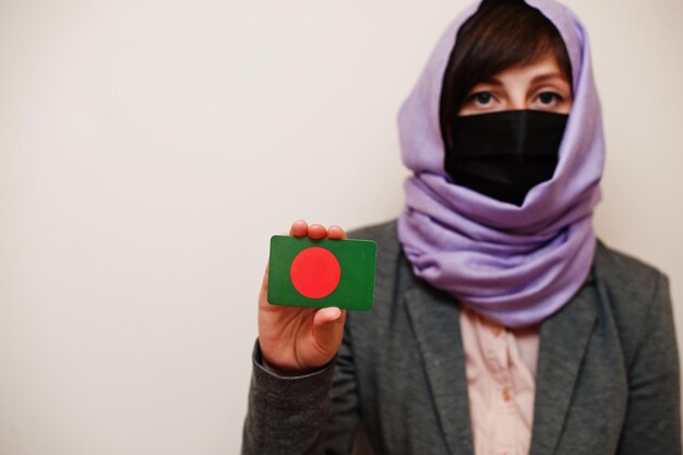 Retrato de una joven musulmana con ropa formal que protege la máscara facial y el pañuelo en la cabeza del hiyab sostienen la tarjeta de la bandera de Bangladesh contra el fondo aislado Concepto de país del coronavirus