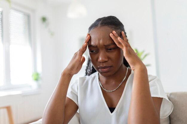 Retrato de una joven negra sentada en el sofá de su casa con dolor de cabeza y dolor de espalda Hermosa mujer que sufre de dolores de cabeza crónicos diarios Mujer triste sosteniendo su cabeza porque tiene dolor en los senos paranasales