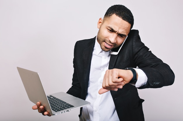 Retrato de joven ocupado trabajador en camisa blanca, chaqueta negra hablando por teléfono y mirando el reloj. Hombre de negocios elegante, trabajando con ordenador portátil, tiempo de trabajo, reunión.