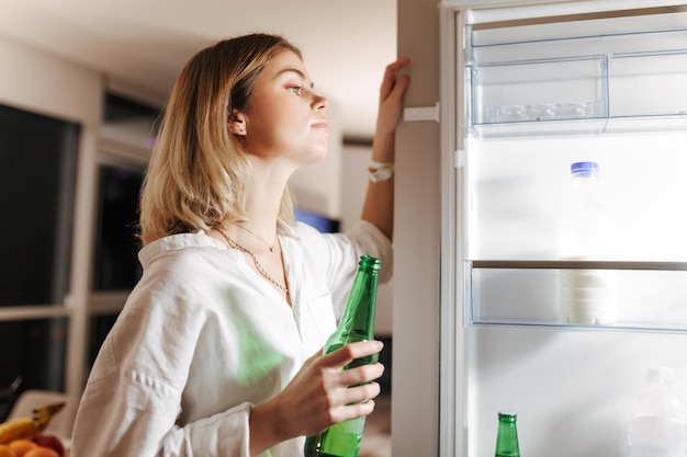 Retrato de una joven parada en la cocina por la noche y mirando soñadoramente en una nevera abierta mientras sostenía cerveza en la mano en casa