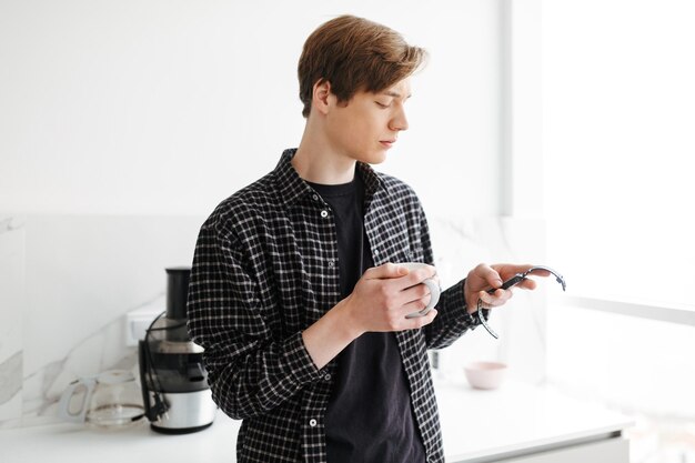 Retrato de un joven parado con una taza en la mano y mirando cuidadosamente el reloj de mano en la cocina de casa