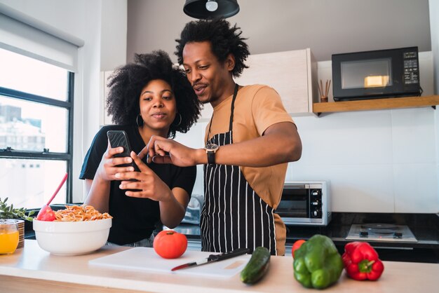 Retrato de joven pareja afro cocinando juntos y usando el teléfono móvil en la cocina de casa.