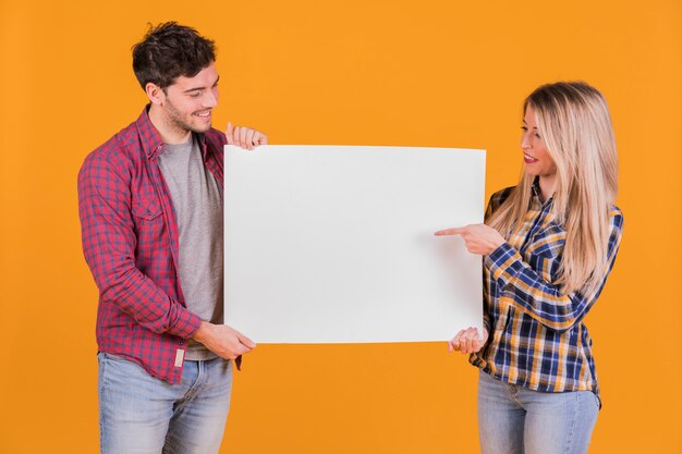 Retrato de una joven pareja apuntando sus dedos en el cartel blanco sobre un fondo naranja