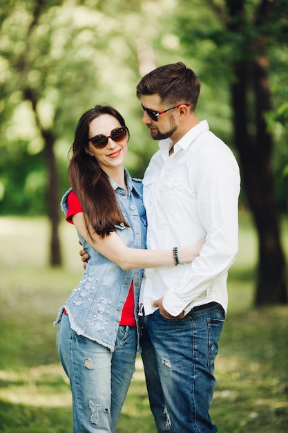 Retrato de una joven pareja feliz enamorada sonriendo y abrazándose en el jardín Dulces amantes con elegantes gafas de sol oscuras y ropa informal posando y mirando la cámara durante el día en el parque de verano