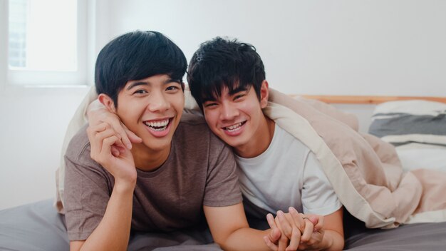 Retrato joven pareja gay asiática sentirse feliz en casa. Los hombres de Asia LGBTQ + se relajan con una sonrisa cariñosa mirando a la cámara mientras descansan juntos y pasan un momento romántico después de despertarse en la habitación de la casa moderna por la mañana.
