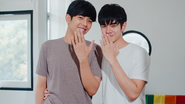 Retrato joven pareja gay asiática sentirse feliz mostrando anillo en casa. Los hombres LGBTQ + de Asia se relajan con una gran sonrisa mirando a la cámara mientras se abrazan en la moderna sala de estar de la casa por la mañana.